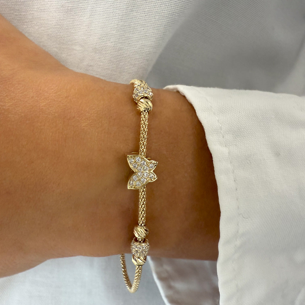 Nahaufnahme eines eleganten Goldarmbands (585/14Karat) am Handgelenk, mit funkelndem Schmetterlingmotiv und Zirkonia-besetzten Pavé-Kugeln, ergänzt durch einen Herzanhänger und Zirkonia Detail an der Verlängerungskette